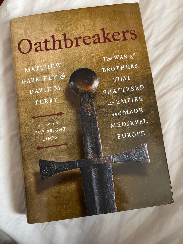 The front cover of Oathbreakers which has a picture of an old metal sword hilt on a dull yellow background with Oathbreakers written in red above. In white text it says The War of brothers that shattered an Empire and made Medieval Europe. It is sitting on my bed (just seen). 