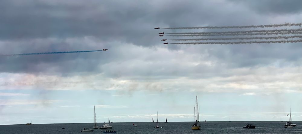 Photo of planes flying across the sky in formation right to left.  One plane is flying towards them.  Grey sea below and grey clouds overhead.  