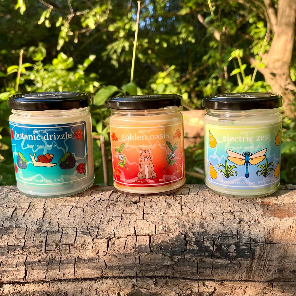 Three candles on a log with foliage behind them. From left to right they are described as follows:

A jarred candle with a black lid and a blue gradient label with an illustration of a snail surrounded by mossy rocks and roses. 
The label reads:

saint fax
botanic drizzle 
moss | rose | earth
hand poured | vegan | 8oz | 100% soy wax


A jarred candle with a black lid and an orange/yellow gradient label with an illustration of a hyena surrounded by amber and sandalwood blossoms
The label reads:

saint fax
golden oasis
amber | sandalwood | musk
hand poured | vegan | 8oz | 100% soy wax

A jarred candle with a black lid and a yellow/blue gradient label with an illustration of a dragonfly surrounded by pears and citronella. 
The label reads:

saint fax
electric zest
pear | citronella | citrus
hand poured | vegan | 8oz | 100% soy wax