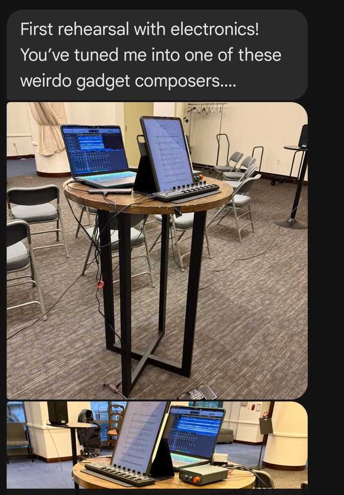 Alt text: A tall round table is set up for an electronic music rehearsal. On the table are a laptop displaying a digital audio workstation (DAW) interface, a tablet showing musical notation, a small MIDI controller with knobs, and an audio interface. Several chairs are arranged in a semi-circle nearby, suggesting a rehearsal space. Cables run from the table to the floor, connecting the devices. The caption reads: “First rehearsal with electronics! You’ve tuned me into one of these weirdo gadget composers….”

