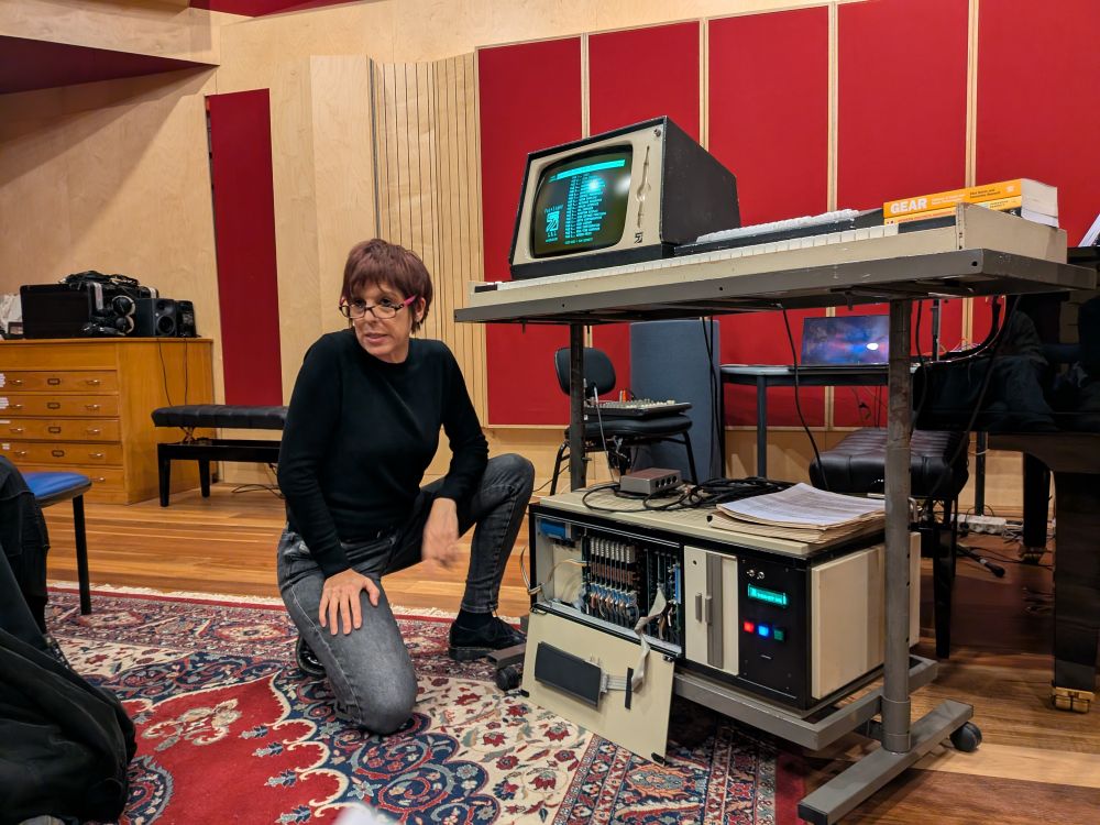 NIME. Building a sequence in real time!! Orch 5 library.

The image shows a woman kneeling on a patterned rug in a recording studio. She is wearing glasses, a black top, and gray jeans. In front of her is a vintage computer music workstation setup, including:

A large CRT monitor displaying a green monochrome screen with what appears to be text and interface elements, possibly running the fairlight software.

An open rack-mounted unit below the table containing 8 units for polyphony.

A QWERTY keyboard and some paper documents on top of the workstation.

The setting has acoustic treatment on the walls and includes other studio gear like a mixer, speakers, and a piano in the background
