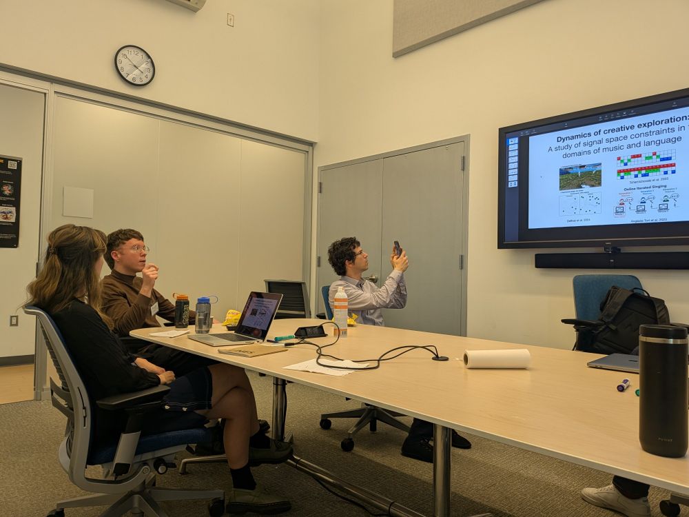 Alt text:
A group of four people is seated around a conference table in a modern meeting room. One person is presenting, gesturing while speaking, while another person takes a photo of a presentation slide displayed on a large screen. The slide is titled "Dynamics of creative exploration" and features graphics related to signal space constraints in music and language. Laptops, water bottles, and a notepad are visible on the table. The atmosphere is focused and academic.

