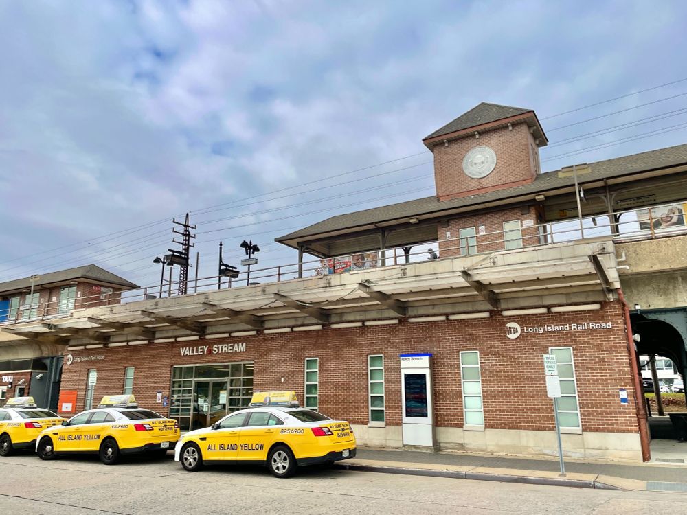A modern-day photograph of Valley Stream station, featuring the station house and the platform overhead. Three taxis are lined up outside the station on a cloudy day.