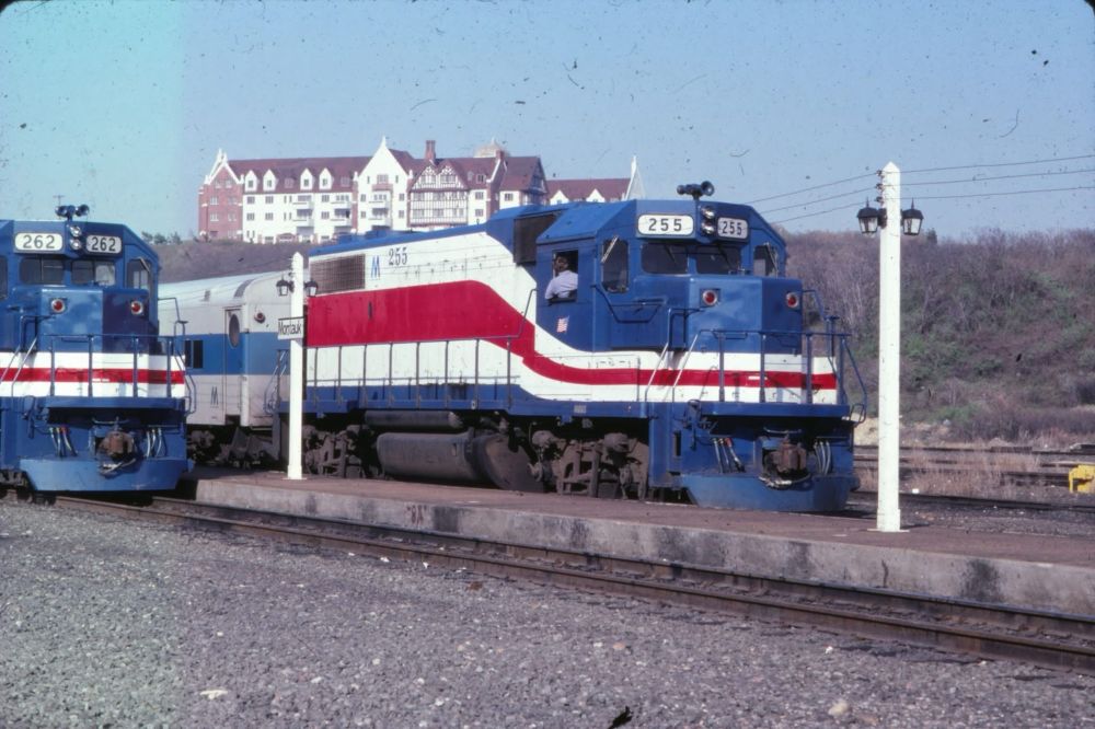 A vintage photo of a red, white, and blue LIRR train running along the tracks on a sunny day in Montauk. 