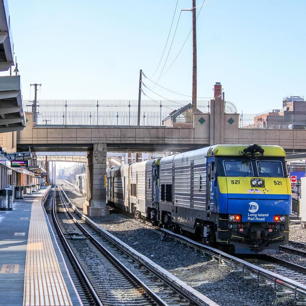 An LIRR diesel train passing by Mineola station on the middle track on a sunny day.