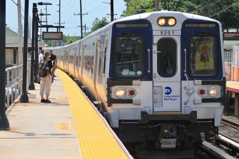 An LIRR M9 train pulling into Cedarhurst station on a sunny day, with people waiting on the platform.