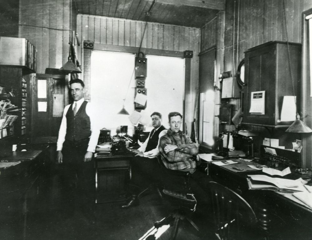 A black and white photograph of a man standing and two seated in the Central Islip ticket office.