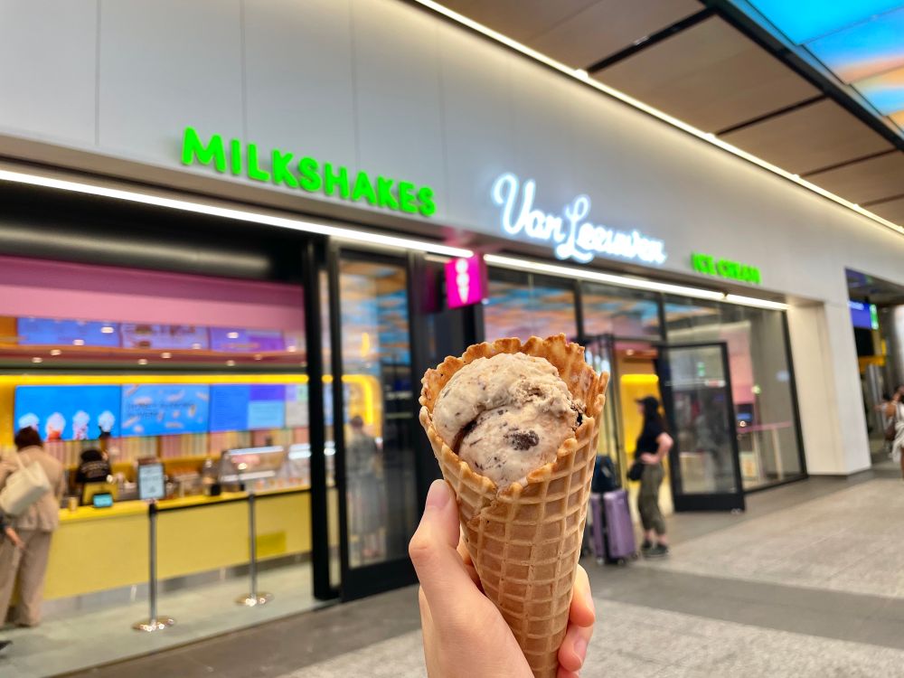 A hand holding up a cone of ice cream, in front of the Van Leeuwen storefront. 