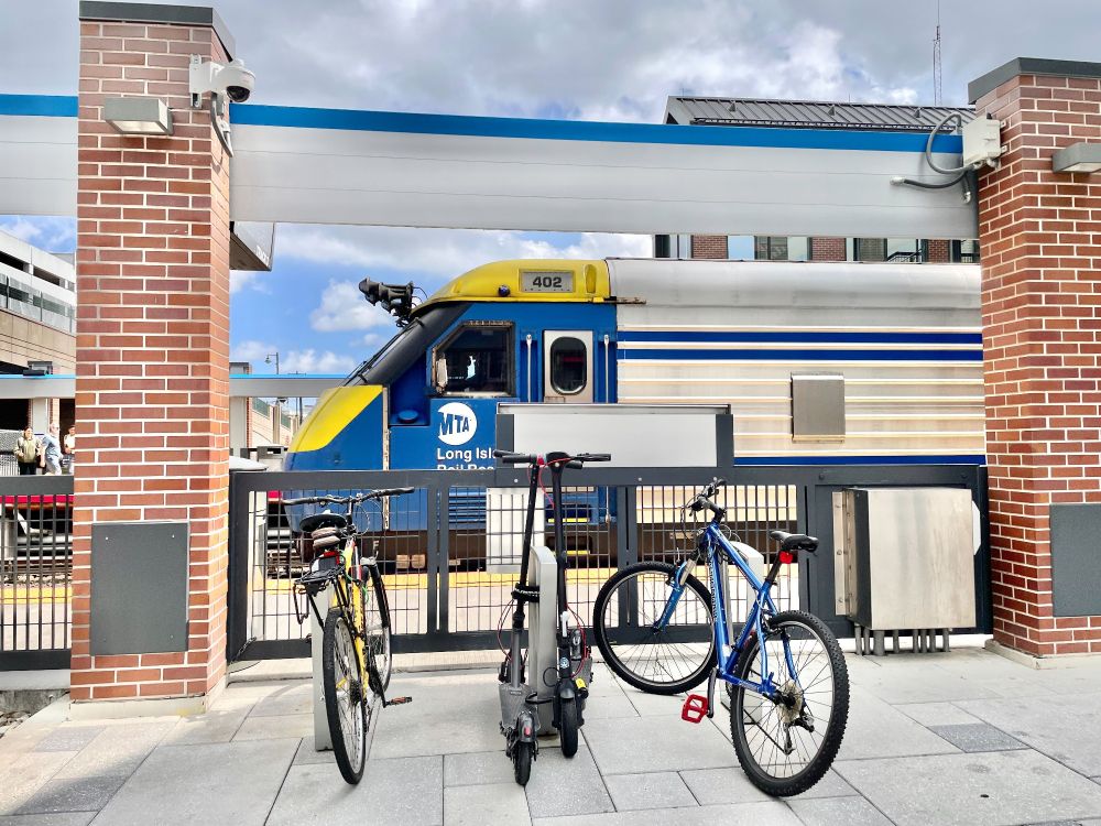 Three bikes at their racks at Mineola station on a sunny day, with a view of an LIRR diesel train passing by in the background.