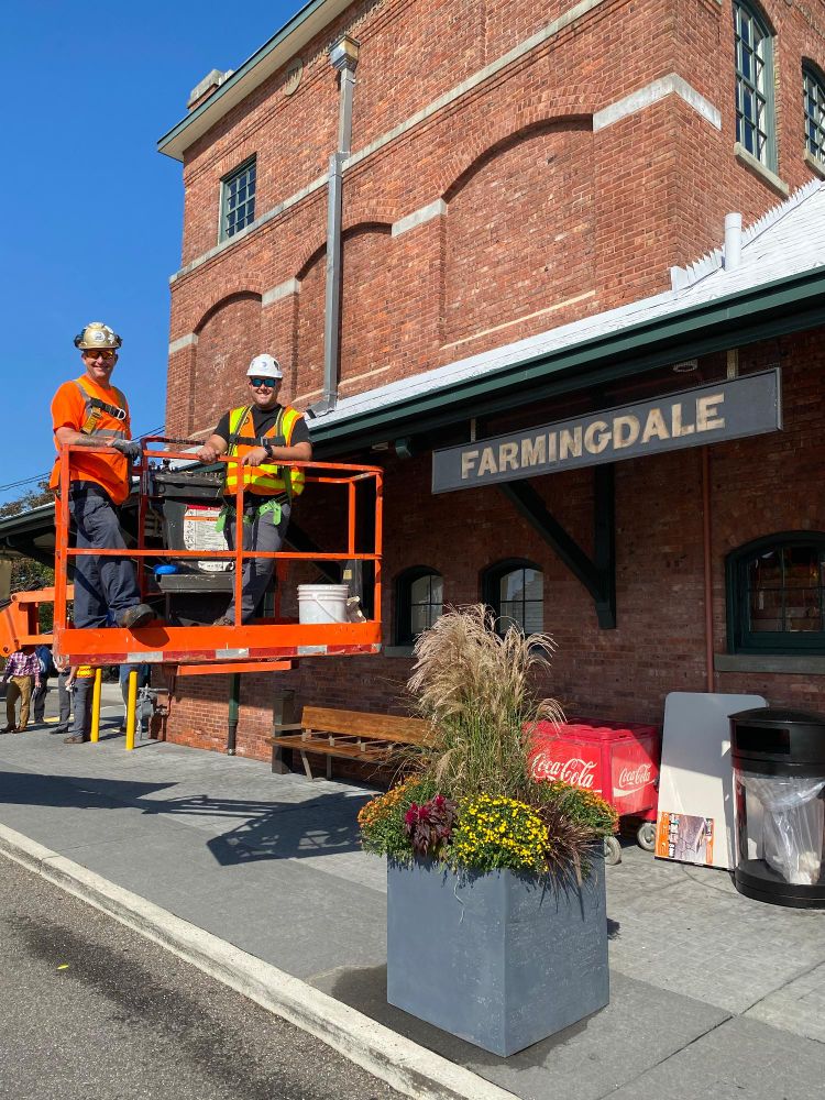 Two LIRR employees wearing helmets and safety vests aboard an orange crane, next to the Farmingdale station building on a sunny day.