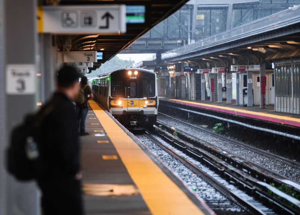 An LIRR train approaching Jamaica station, its lights flashing, with people waiting on the platform on a rainy day.