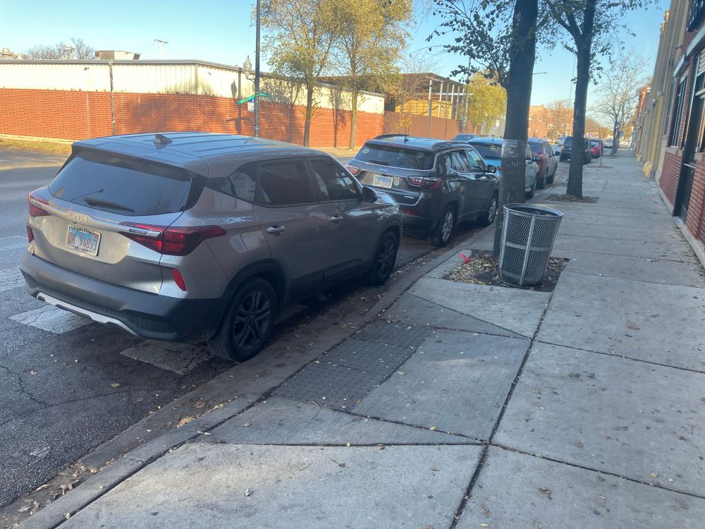 2 cars parked blocking crosswalks. 1 in the foreground, one visible in the background with cars parked between them.