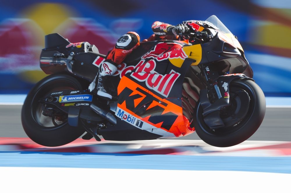 Pedro Acosta, riding a blue and orange KTM RC16 MotoGP bike, on the exit of Turn 16 at Misano. He is riding the outside of the kerbs, with both wheels off the ground, and it looks like he is flying. Probably because he is. 

Behind him is a blurred Red Bull logo.