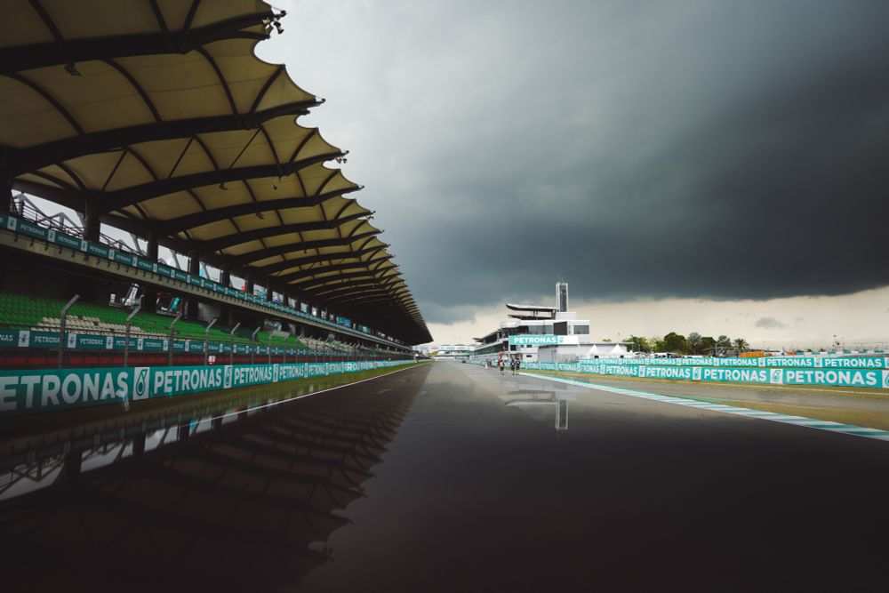 A photo of the front straight at Sepang. On the left is a grandstand, on the right in the distance is the pits. Dark clouds hang over the whole scene, and there are faint reflections in the bottom half of the photo