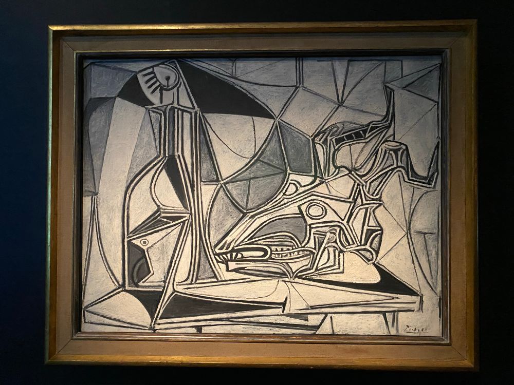 A grayscale cubist painting is displayed in a gold frame. The painting features various geometric shapes and fragmented forms that coalesce into abstract representations of objects. On the left, a tall, slender form with an eye-like shape at the top could be interpreted as a stylized figure or bottle. To the right, a more complex arrangement of lines and curves suggests an animalistic or mythological creature, possibly a horse or a griffin, with a prominent head and elongated snout or beak. The background is composed of intersecting lines and planes, characteristic of cubist art, creating a sense of depth and fractured perspective. The color palette is restricted to shades of black, white, and gray, enhancing the stark, geometric nature of the composition. The artist's signature, "Marcel," is visible in the bottom right corner.
