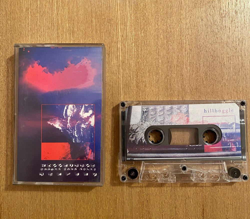 The image shows a cassette tape and its case placed on a wooden surface. The case is on the left side and features abstract artwork with a mix of dark blue, purple, and red colors, with a square section in the center containing a distorted, high-contrast image. Below the artwork, there is stylized, almost unreadable text or symbols. The cassette tape is on the right side, housed in a transparent plastic shell. The label on the cassette displays a collage-style design with muted colors and the word “hillboggle” written in small, delicate text in the top right corner. The cassette tape spools and mechanisms are visible through the clear plastic.