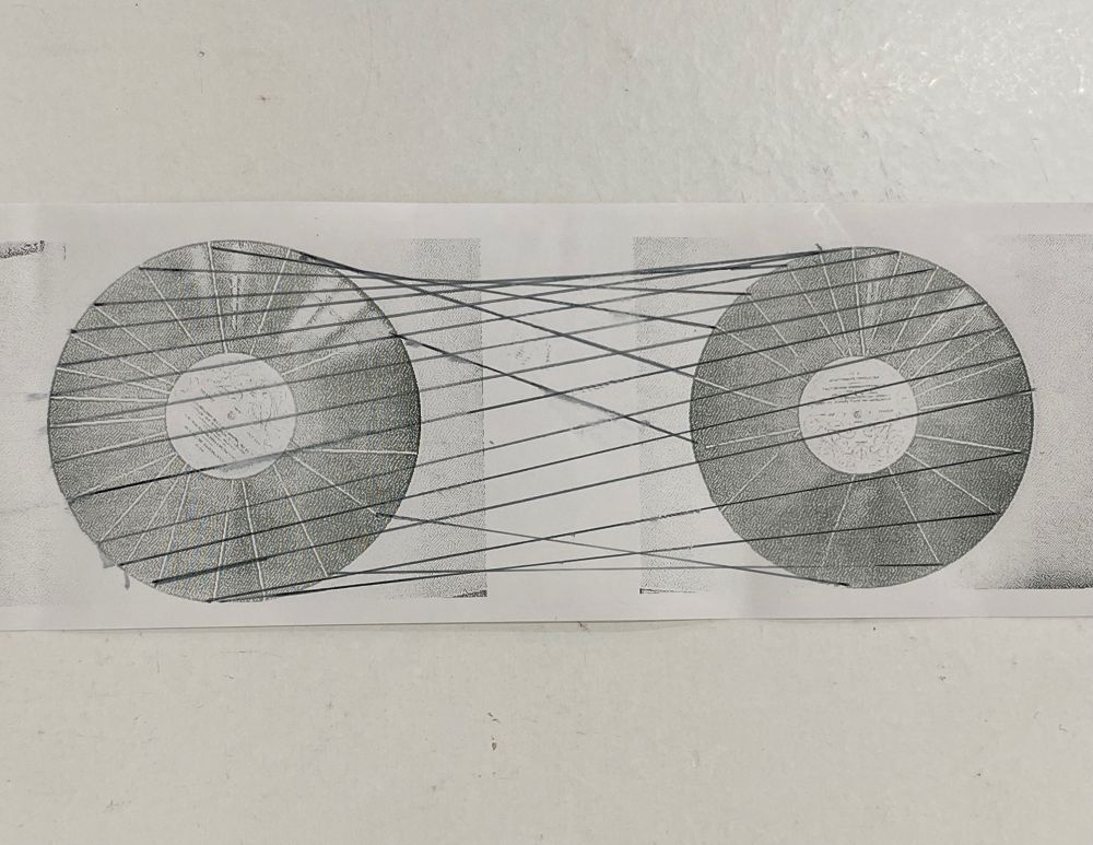 A long, rectangular, black and white image shows two identical, circular objects, resembling phonograph records or spools, placed side by side. The circles have a central label and a pattern of lines radiating outwards, intersected by concentric circles. A series of straight, dark lines are drawn between the two circles, connecting points on the circumference of one circle to points on the circumference of the other. The lines cross over each other in the space between the circles, forming a complex, crisscross pattern. The image is a print on a light-colored surface, possibly paper, with the top and bottom edges slightly faded or torn.
