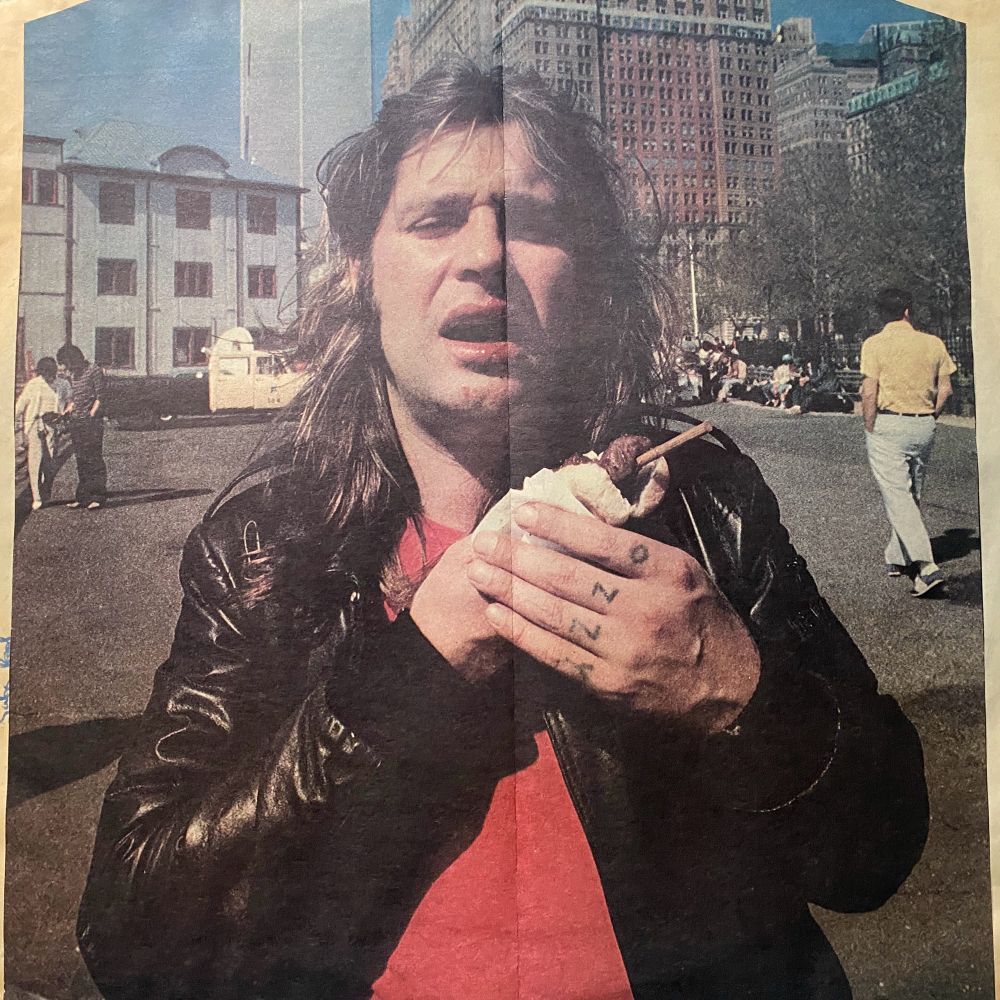A man with long, wavy hair and a black leather jacket is standing outdoors in a city park, holding an ice cream cone with a chocolate topping. He is wearing a red shirt underneath the jacket. The fingers on his right hand are visible and display the tattooed letters “OZZY.” Behind him, people are sitting and walking in the sunny park, and tall buildings line the background. The photo has a vintage quality and appears to be from the late 1970s or early 1980s.