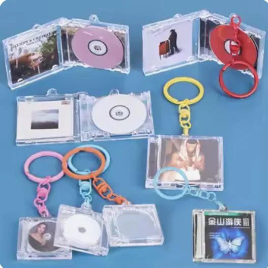 A top-down shot showcases nine mini CD album keychains arranged on a blue surface. Each keychain features a clear, square plastic case, mimicking a real CD album, with various designs. Some cases are open, revealing a miniature CD in a different color (like pink, white, or red) on one side and album art on the other. Other cases are closed, displaying the album art. Each case is attached to a colorful plastic keyring in shades of pink, orange, yellow, blue, or red.
