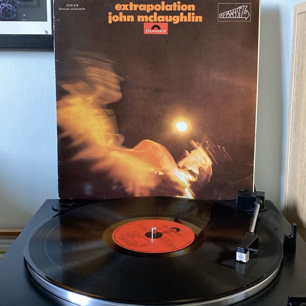 A vinyl record of the album "Extrapolation" by John McLaughlin is playing on a turntable. The record is black with an orange label. The album cover features a blurry, motion-filled image of John McLaughlin playing guitar, illuminated by a single light source. The cover has the artist's name and album title in orange text at the top.
