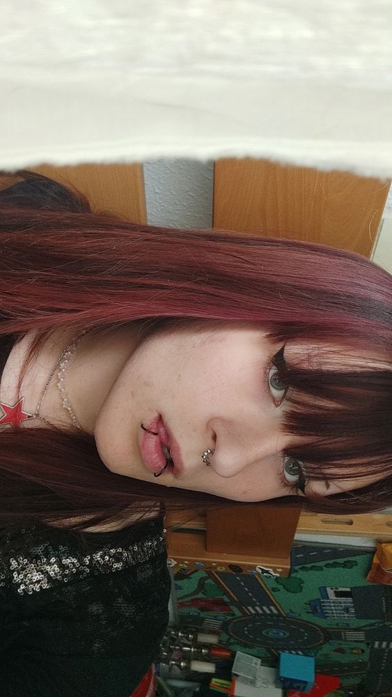 Girl showing her face while having a cheeky look. She's wearing sleek eyeliner with a lot of accessories. Her hair is long, brown-red.