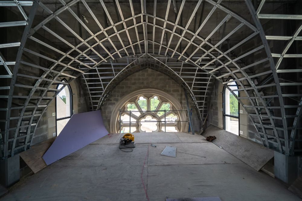 Vault framework looking the W rose window. Notice how there are studs which attach to the concrete roof. To the L is a piece of cut purplish panel which is presumably the surface that is applied to the studs. There is also an electric handsaw on the wooden work platform.