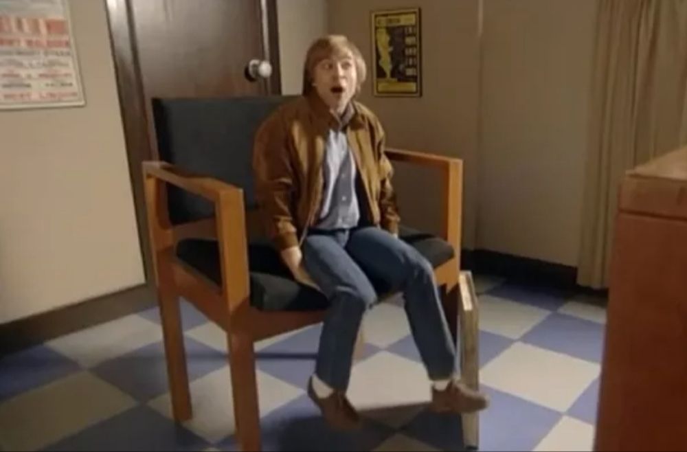 A still from there 'Little Britain' sketch where David Walliams is playing the character of Dennis Waterman. He's sitting on an oversized chair in a scaled up office setting to make him look the size of a child in the sketch. The catchphrase for this sketch was "I'm going sing the 'feem' tune, I'm going to write the 'feem' tune"
