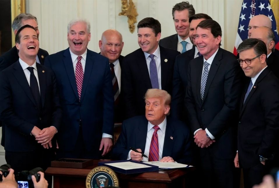 A photo of ten men in suits smiling and laughing, gathered around the back of Donald Trump's chair. Donald Trump is sitting there, holding a pen above some sheets of paper, unsmiling and looking as far through as a clear day. 
