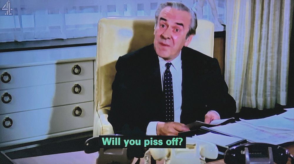 John Le Mesurier sitting at a desk in the middle of saying something to someone out of frame. The subtitle says "Will you piss off?"