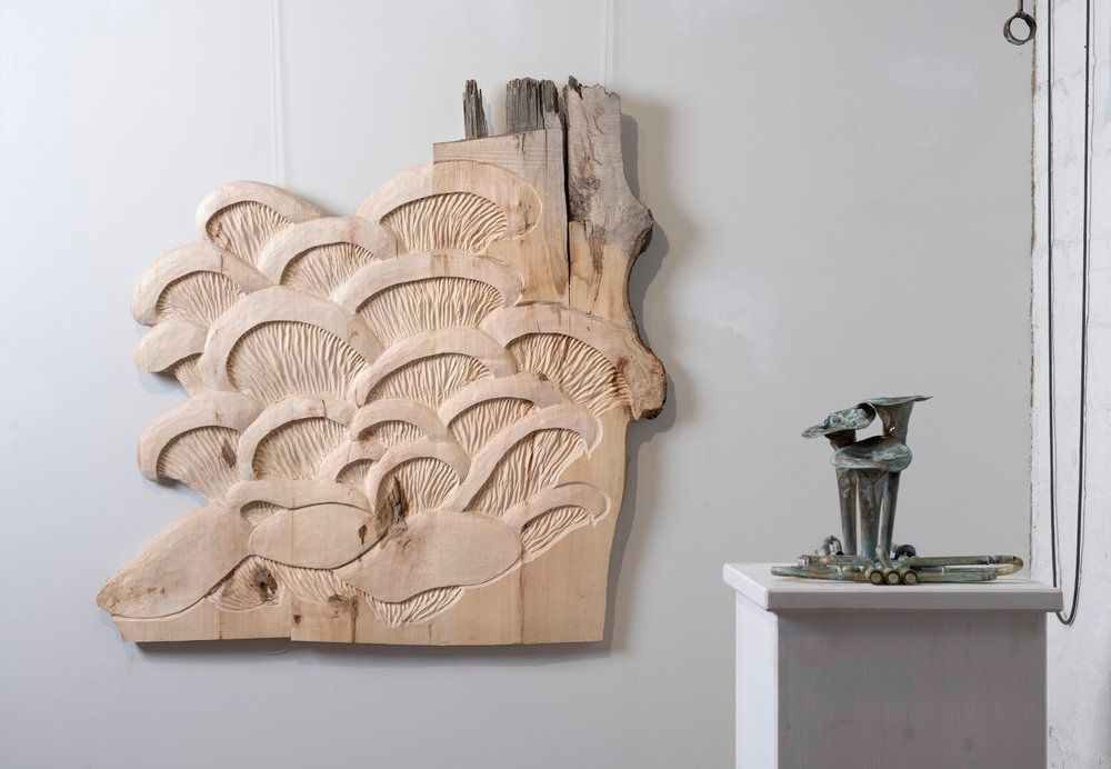 Wood carving of oyster mushrooms on a tree stump in a Gallery space with a bronze sculpture on the side.