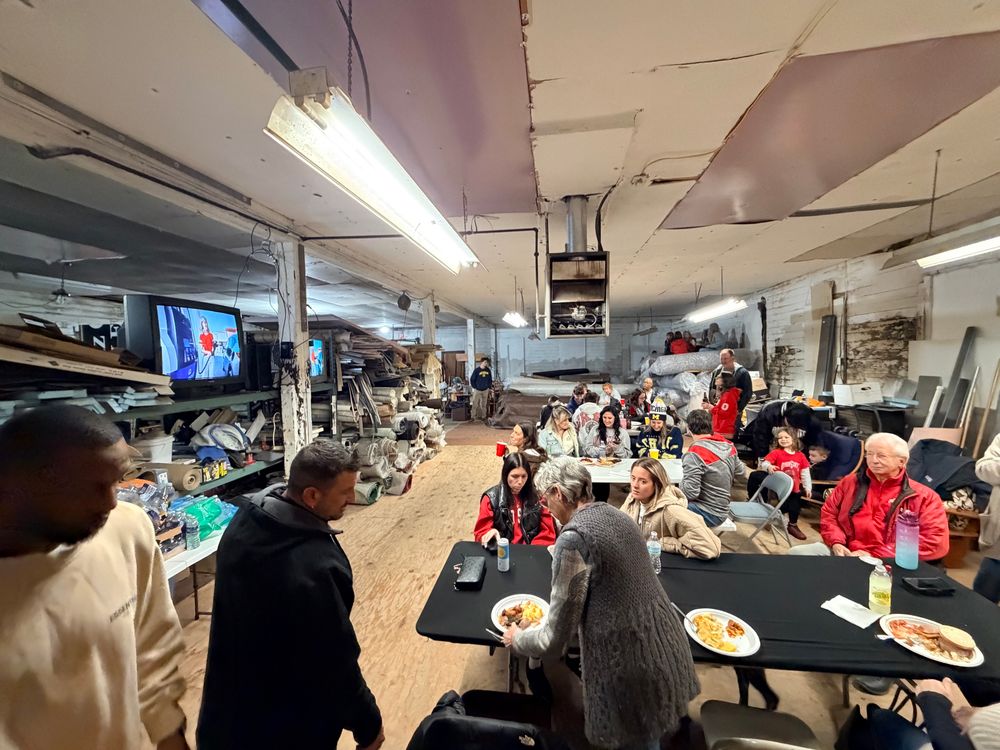 A large group of people gathered for a meal inside an old, cluttered workshop or warehouse. Long folding tables and plastic chairs are set up, with plates of food and drinks in front of guests. The room has exposed beams, mismatched ceiling panels, and industrial fluorescent lights. Shelves along the left wall are stacked with rolled-up carpets, tools, boxes, and miscellaneous materials. Two TVs mounted on the wall display the game. People of various ages—kids, adults, and seniors—sit and stand in small groups, talking and eating. Some wear winter coats and hats, giving the scene a casual, makeshift holiday gathering atmosphere.