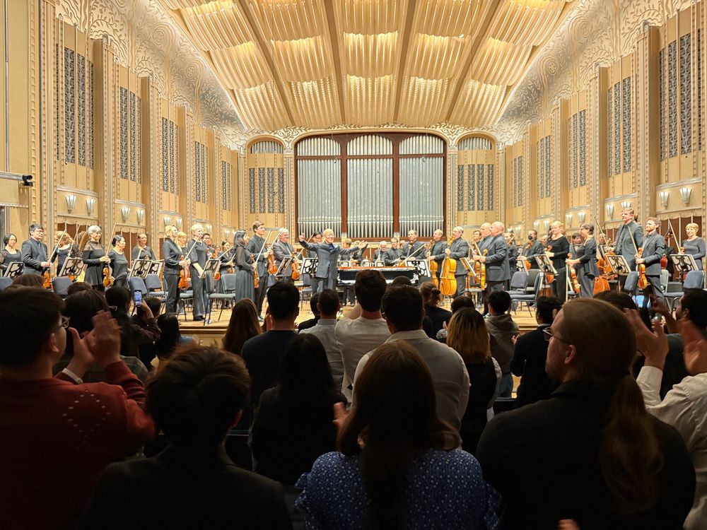 Audience in a concert hall giving a standing ovation to the Cleveland Orchestra, with musicians standing on stage holding their instruments and the conductor taking a bow. The ornate hall features golden lighting, carved details, and a large pipe organ as the backdrop.