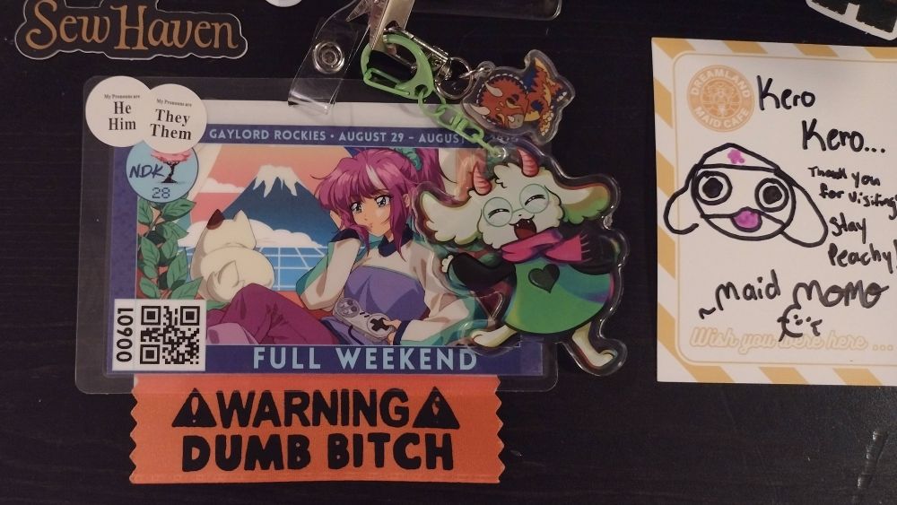 a charm of Ralsei from Deltarune, a charm of a Triceratops and an orange ribbon with the text "Warning Dumb Bitch" all on my NKD Full Weekend 2025 badge 
