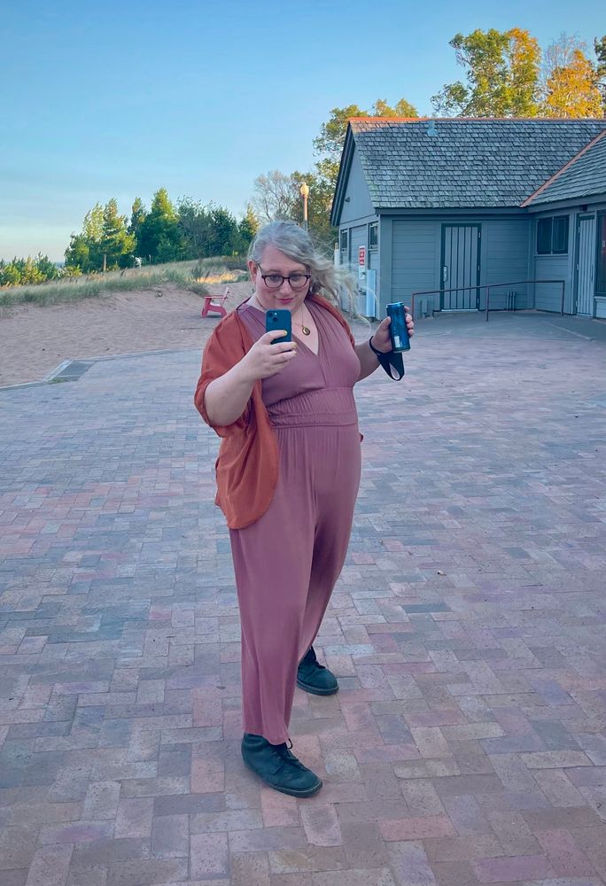 Suswave poses in a mauve jumpsuit with a drapey orange thing. they're on a brick patio next to the beach. they're taking a picture of the picture-taker. they look satisfied. their tits are awesome. sorry. I just. it had to be said