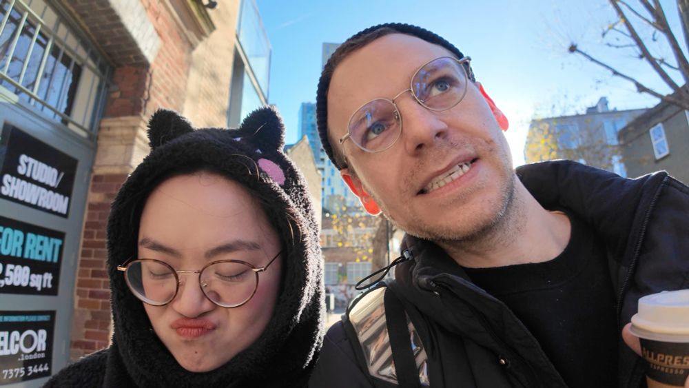 Double portrait of Yoyo and David talking to camera as they walk through the streets of London. Both are wearing black, coats, clothes and hats, with Yoyo wearing her cat hood with ears that pop up.

The shot is taken towards the sun, with David’s heard to the right of the image obscuring the sun location, Yoyo’s head to the left of the image is expressive, pouting and looking downwards, seemingly g in response to something David is sharing. This shot is taken from an episode of the vlog currently available on YouTube. 