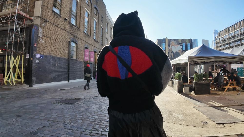 A central composed photo of Yoyo, dressed in black with a big red eye graphic in her back, facing the camera. We do not see Yoyo as she is facing away, looking out towards a courtyard in London, UK. The environment is a socia areal with tents and seating, next to a location called ‘Truman’s brewery’. Light it shining over the top of a large factory building on the left, into the courtyard. With blue skies above. Yoyo is wearing mostly black, including a black cat hood, which has ears poking out to create a unique profile.