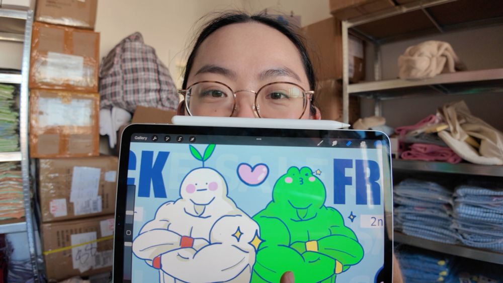 A medium shot/close up of Yoyo sitting in from of the camera, inside the studio. Yoyo is holding her iPad up to the camera to show her artwork for a holiday sale social media post. The iPad partly obscures her face as it it in from of her face, showing only the ridge of her nose upwards, including eyes, glasses, top of head and hair bordering her face skin.

The background is the studio environment, showing shelving and boxes, product placed on shelves and miscellaneous items places around. 

The image is lit by sunlight, and is warm.
The graphic on the iPad shows two muscular characters standing shoulder to shoulder, with the letter CK and FR either side, indicated the image has been zoomed into.