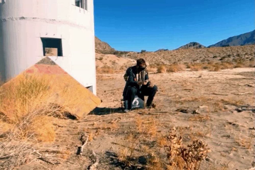 a person in a gas mask sits in the dirt near a pyramid