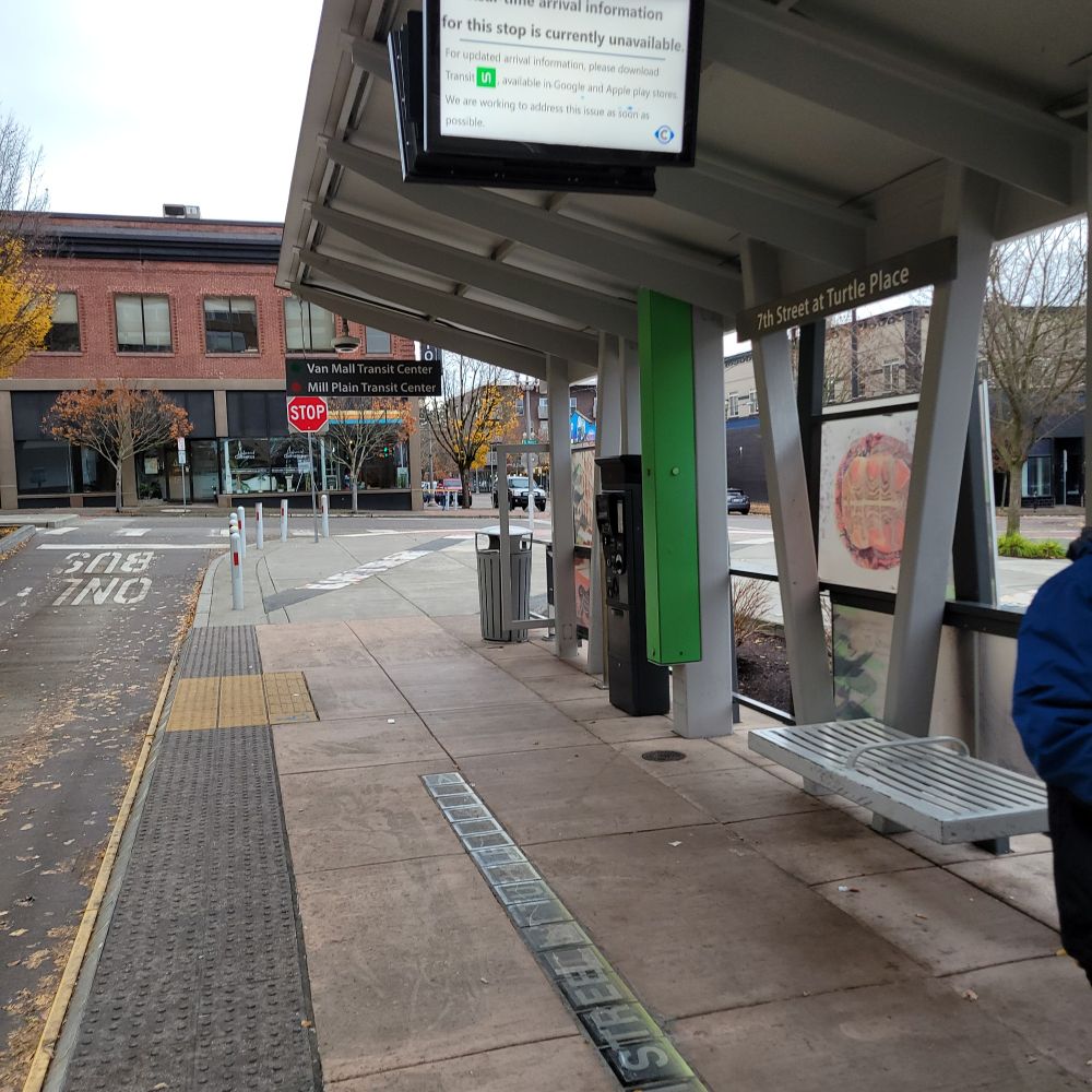 Photo of a transit stop with benches, roof, yellow truncated dome indicator for boarding spot, overhead sign that usually provides transit arrival info but right now directs people to use the Transit app.