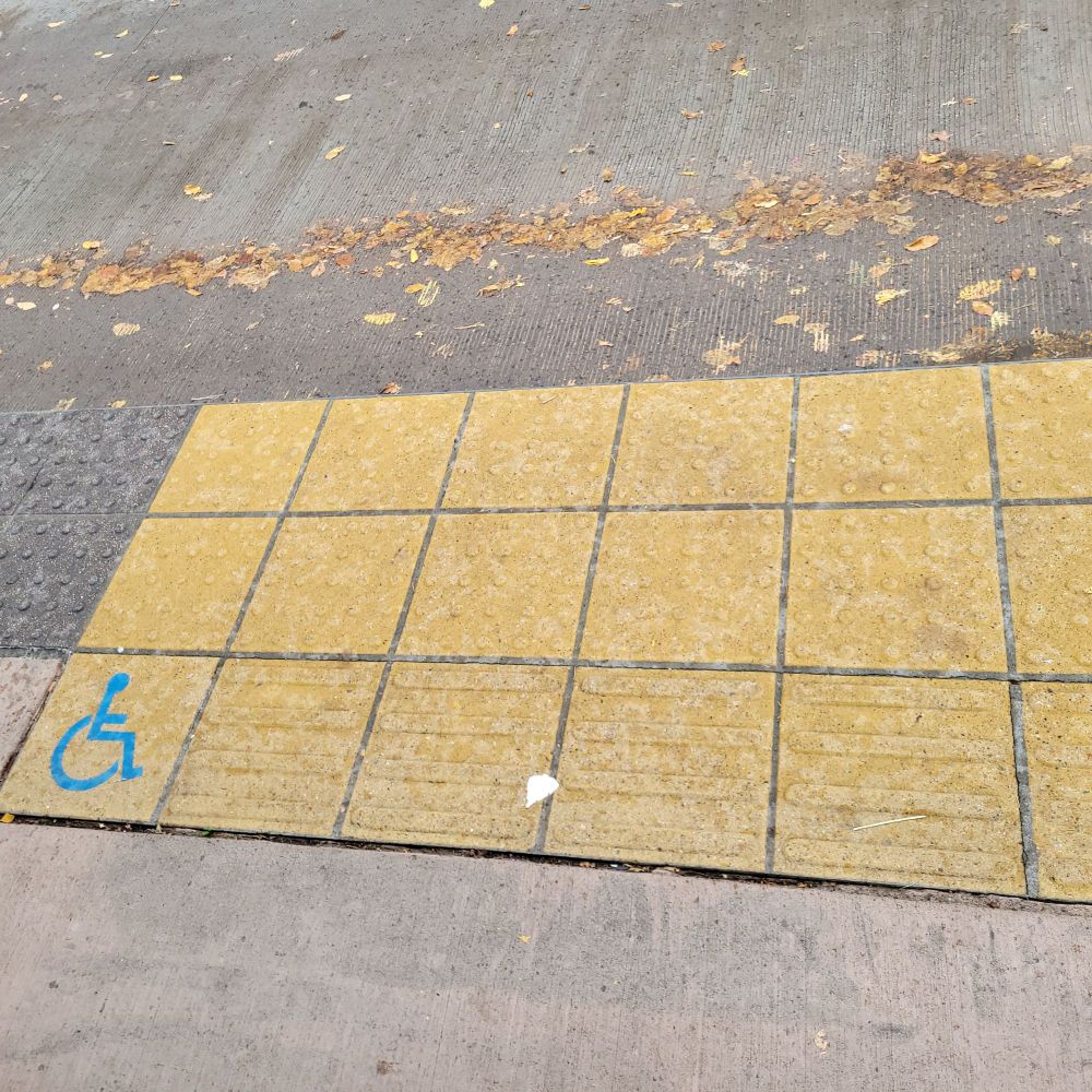 Close up photo of a yellow checkerboard pattern on the sidewalk with a wheelchair user icon in the lower left-hand corner.