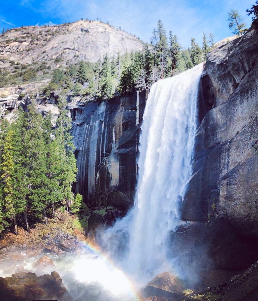 Millions of gallons of water pour over a tall granite slab. Vernal Falls has green trees to the left of it and a tall peak above it.