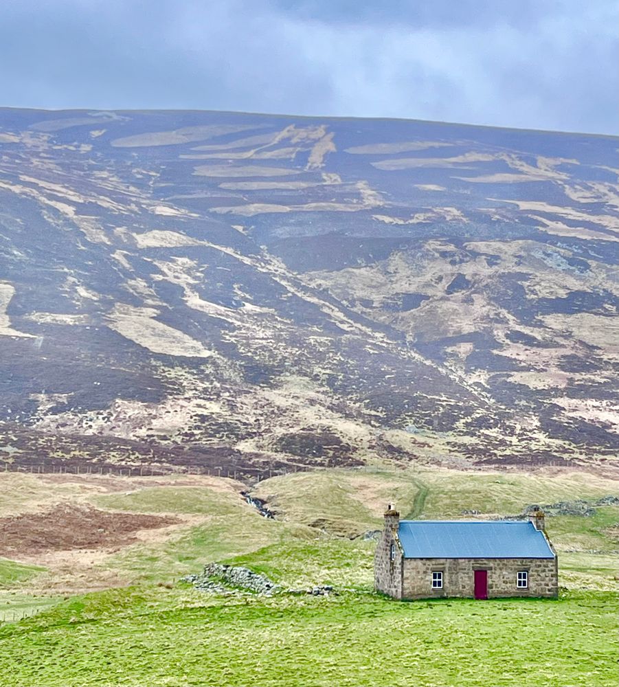 A small stone tan house with a blue roof and a red door sits at the base of the Scottish highland mountains.