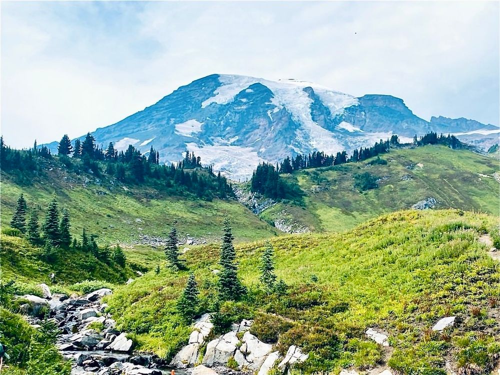 A majestic mountain appears blue in the distance behind a tree topped green ridge line. A small boulder covered brook trickles in the lower left side of the image.