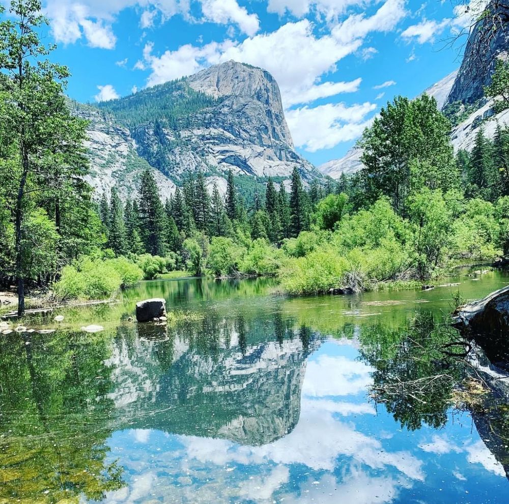 A granite mountain reflects in a pristine still lake. It is surrounded by evergreen trees and there is a blue sky with white clouds above it.