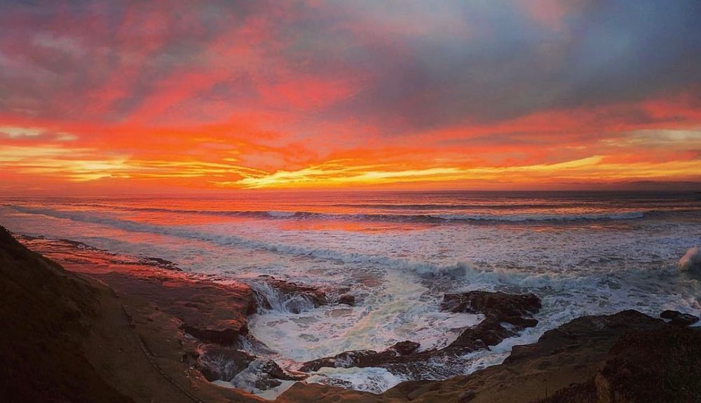 The sky is ablaze in oranges, purples, and yellows as the sun sets over the dark blue ocean with white waves crashing on black boulders in the forefront. 