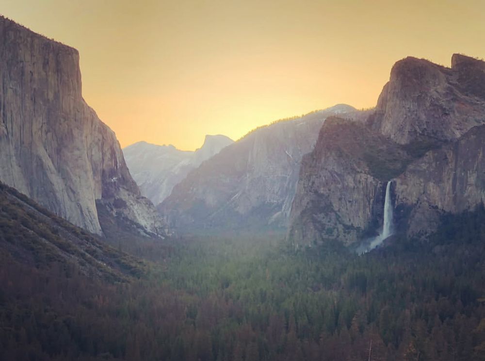 A bright orange sunrise begins to glow over Yosemite Valley. Bridalveil Falls flows to the rights of the picture, while El Capitan towers to the left.