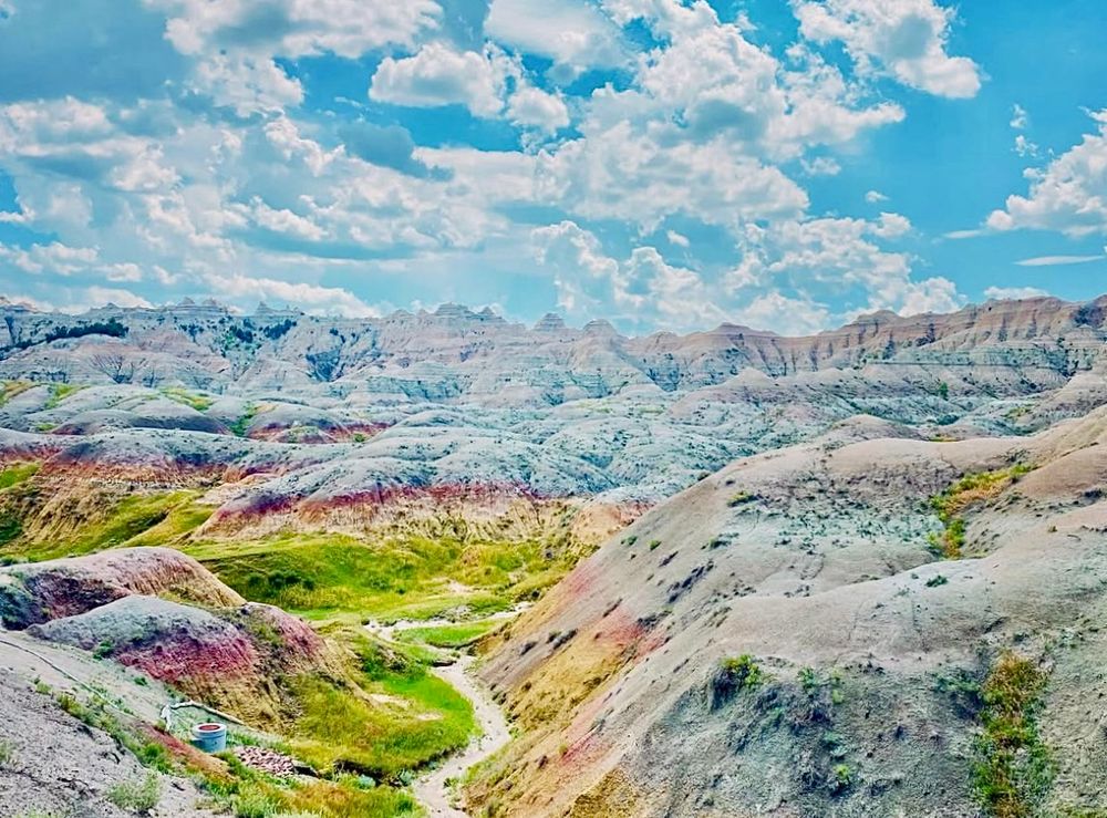 A rugged landscape featuring yellow, pink, and gray rock. A bright blue sky above full of wispy white clouds. Bright green grass at the bottom of the canyon.