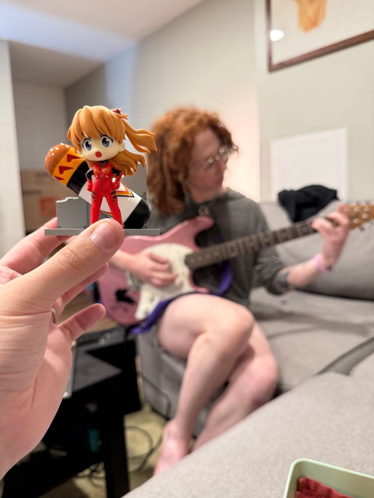 Jordan holding a plastic figure of Asuka from Evangelion, in front of Stella, who is playing her pink electric guitar. They are both redheads 