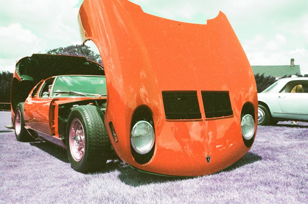 A Lamborghini Miura in red, sits open on a lawn. Due to the film stock used the grass is purple and otherworldly. 