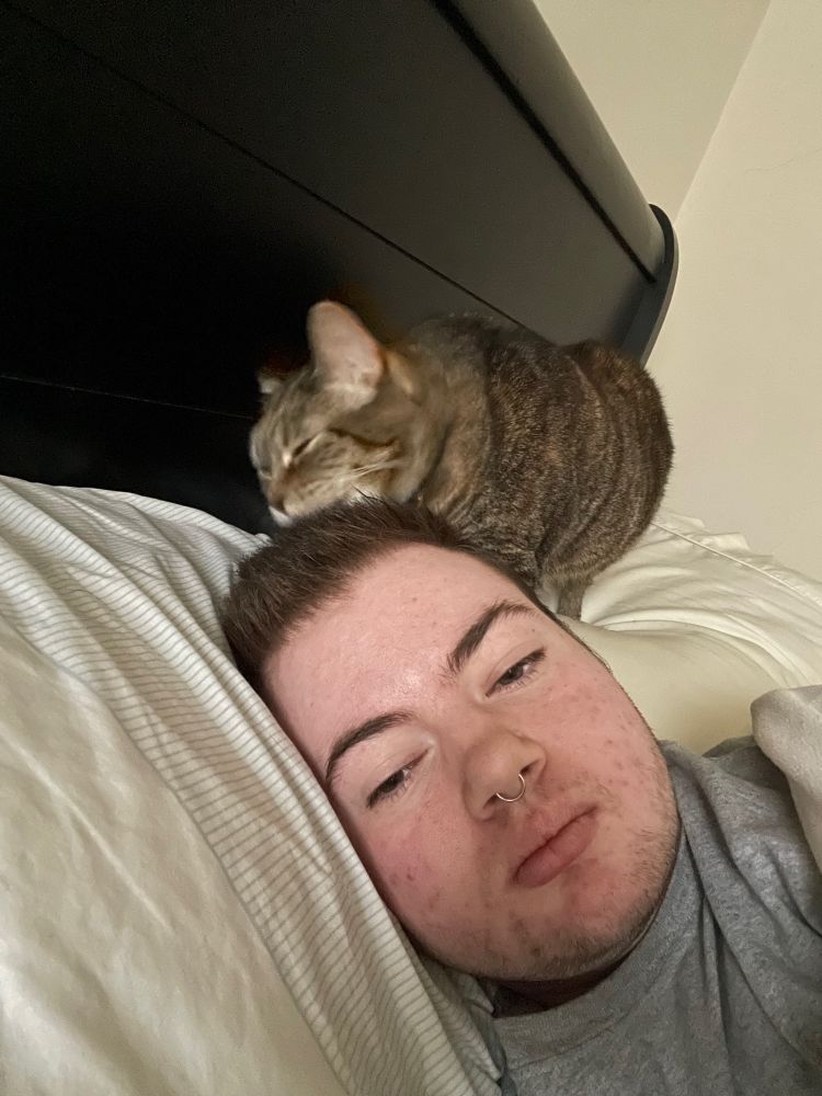 white guy with chubby face and grown out buzzcut is pictured along with a torbie cat licking the guy’s hair